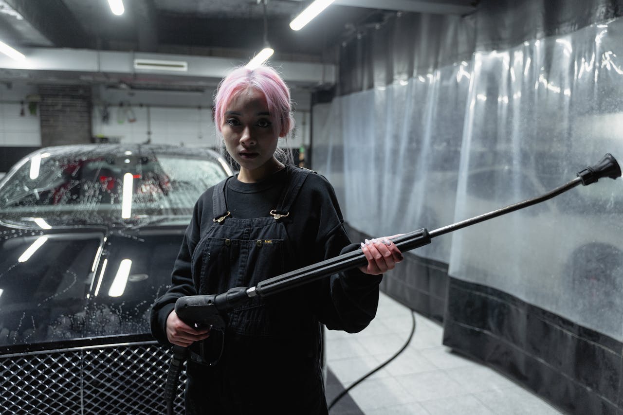 A woman with pink hair uses a pressure washer in a car wash setting. Indoor, nighttime shot.